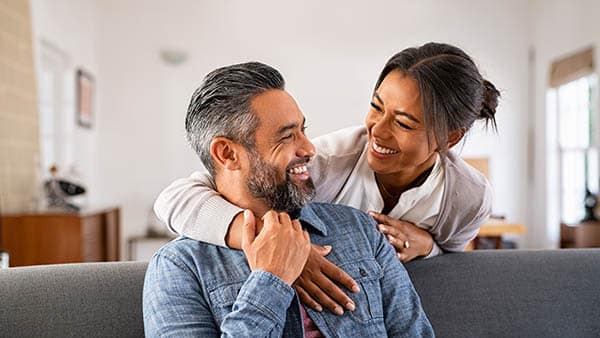 Smiling woman hugging her husband on the couch from behind in the living room.